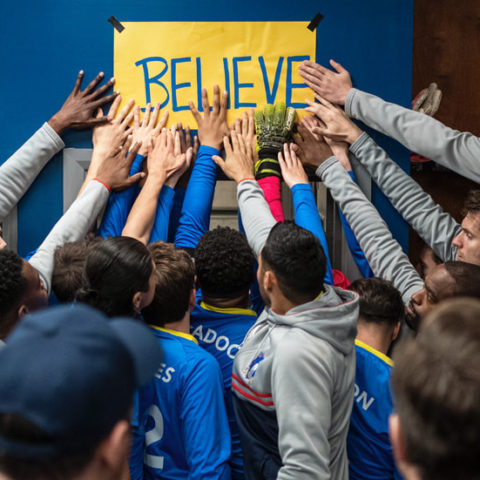 The squad of AFC Richmond from Ted Lasso, place their hands on a sign reading 'Believe'